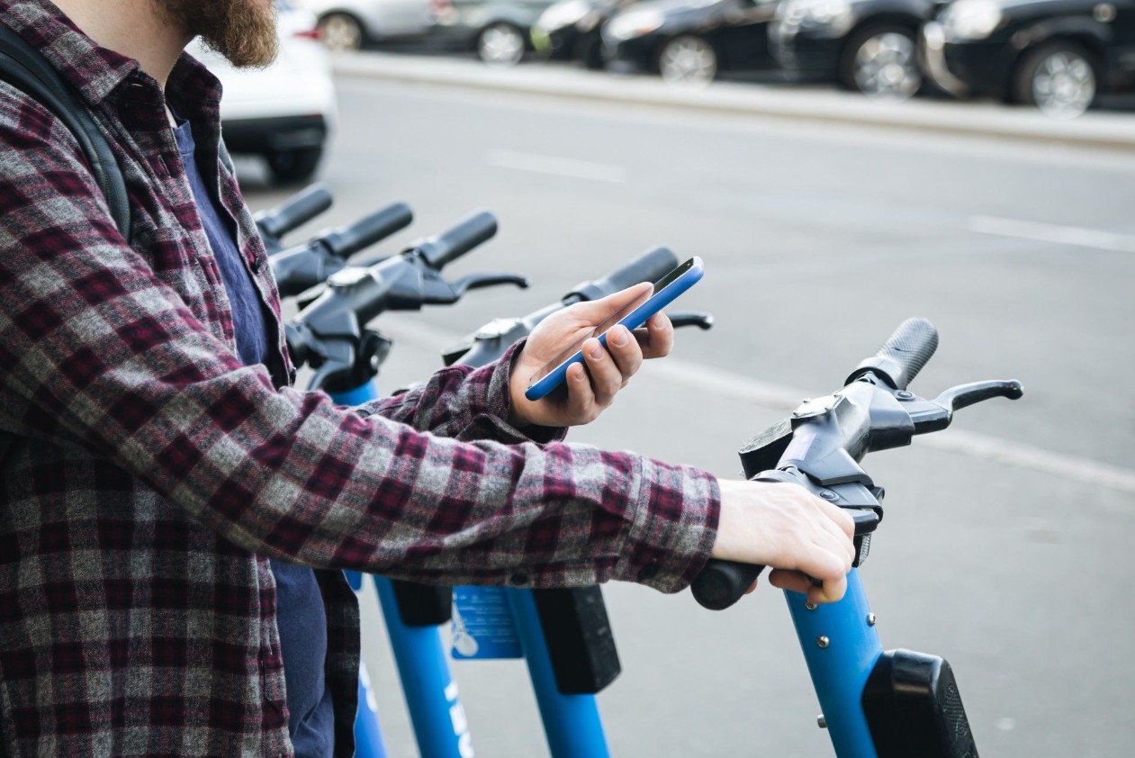 Homme tenant son téléphone dans une main et le guidon d’une trottinette électrique dans l’autre.